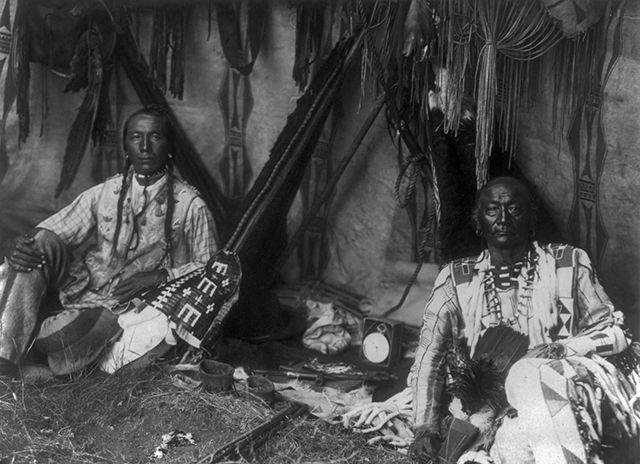 Little Plume and son Yellow Kidney seated on ground inside lodge, pipe between them. Curtis was criticized for removing traces of non-Native culture, like the clock, from his photos.