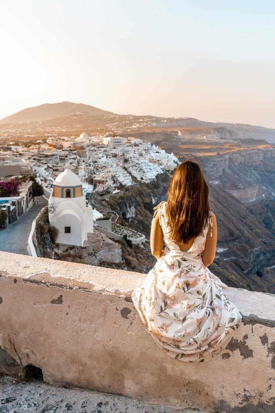 Girl in a floral dress sitting on a wall, watching the sunrise over Fira