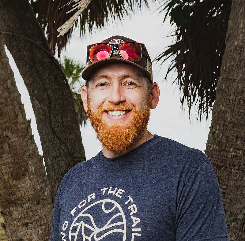Mason Gravely portrait with palm leaves in the background. Mason is waearing a baseball hat with sunglasses on his hat wearing a two for the trails Athletic Brewing Co shirt.
